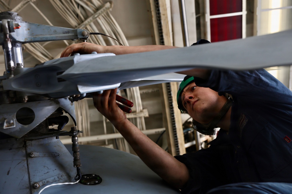 A technician wearing a green cap and dark coveralls is performing maintenance on helicopter rotor blades in what appears to be a hangar bay, likely aboard a naval vessel. The person is closely inspecting or adjusting the rotor hub assembly, leaning in for a detailed examination. The setting and attire suggest a military aviation maintenance environment.