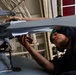 A technician wearing a green cap and dark coveralls is performing maintenance on helicopter rotor blades in what appears to be a hangar bay, likely aboard a naval vessel. The person is closely inspecting or adjusting the rotor hub assembly, leaning in for a detailed examination. The setting and attire suggest a military aviation maintenance environment.