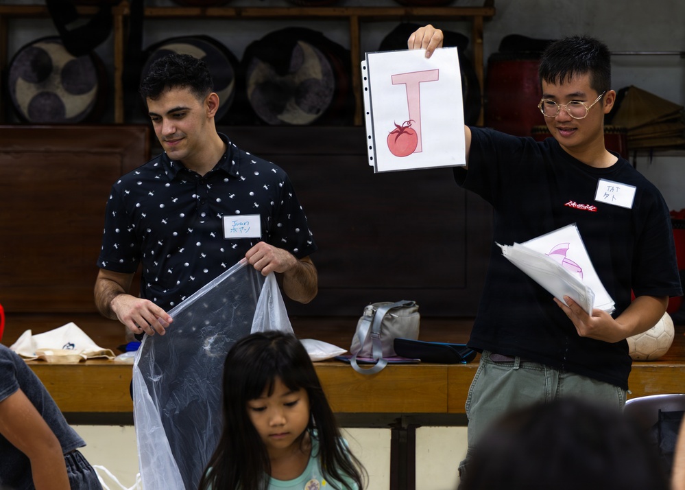 Two young men wearing name tags are presenting educational materials in what appears to be a classroom or community hall setting. The person on the right is holding up a flashcard showing the letter 'T' with a tomato illustration, suggesting an English language learning activity. A young child is visible in the foreground, and traditional-looking drums or instruments are visible in the background.