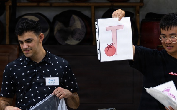 Two young men wearing name tags are presenting educational materials in what appears to be a classroom or community hall setting. The person on the right is holding up a flashcard showing the letter 'T' with a tomato illustration, suggesting an English language learning activity. A young child is visible in the foreground, and traditional-looking drums or instruments are visible in the background.