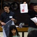 Two young men wearing name tags are presenting educational materials in what appears to be a classroom or community hall setting. The person on the right is holding up a flashcard showing the letter 'T' with a tomato illustration, suggesting an English language learning activity. A young child is visible in the foreground, and traditional-looking drums or instruments are visible in the background.