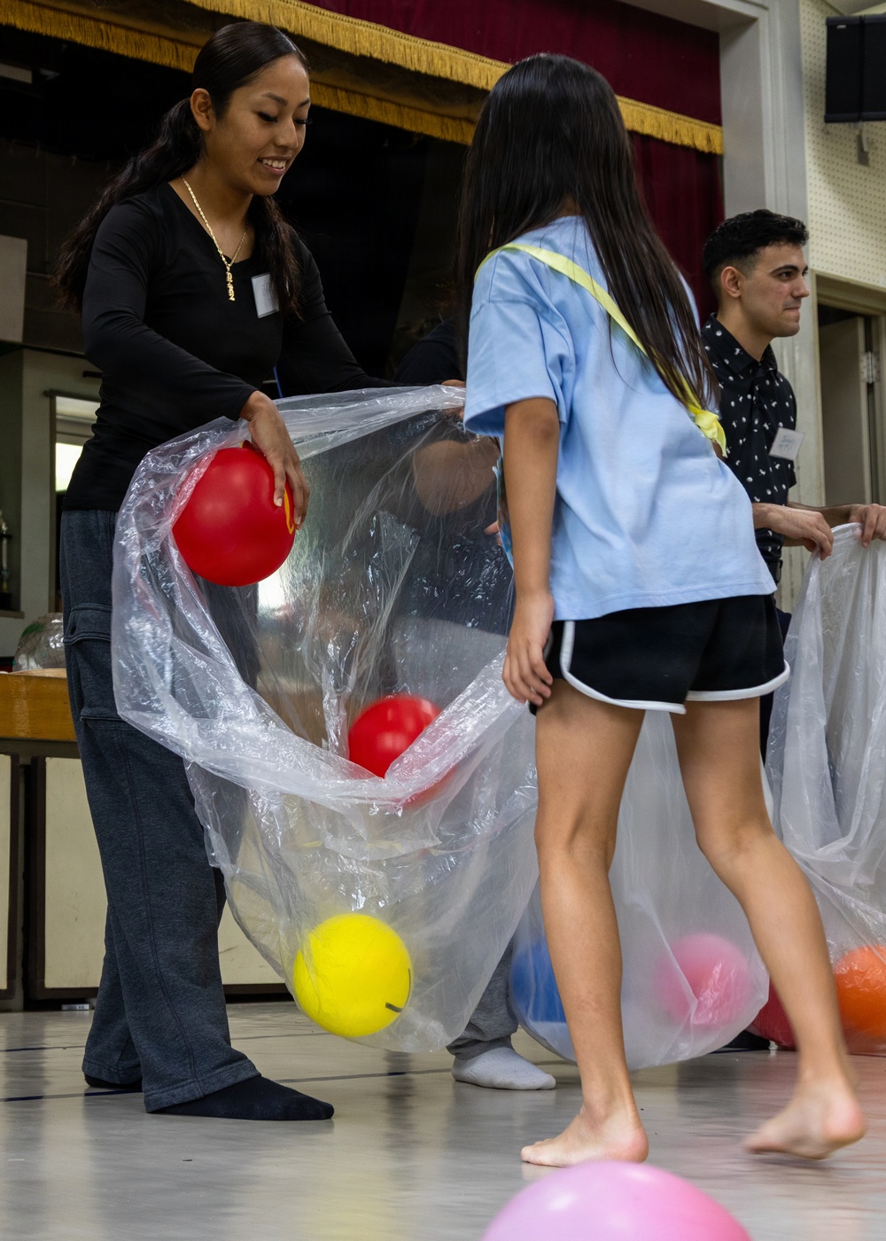 A group of people are engaged in an indoor activity involving large clear plastic bags and colorful balls. An adult woman in a black top and a young girl in a light blue shirt are cooperatively holding a transparent bag containing red and yellow balls. The setting appears to be a hall or gymnasium with a stage backdrop visible in the background.