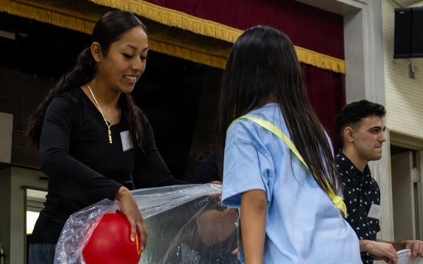 A group of people are engaged in an indoor activity involving large clear plastic bags and colorful balls. An adult woman in a black top and a young girl in a light blue shirt are cooperatively holding a transparent bag containing red and yellow balls. The setting appears to be a hall or gymnasium with a stage backdrop visible in the background.