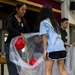 A group of people are engaged in an indoor activity involving large clear plastic bags and colorful balls. An adult woman in a black top and a young girl in a light blue shirt are cooperatively holding a transparent bag containing red and yellow balls. The setting appears to be a hall or gymnasium with a stage backdrop visible in the background.