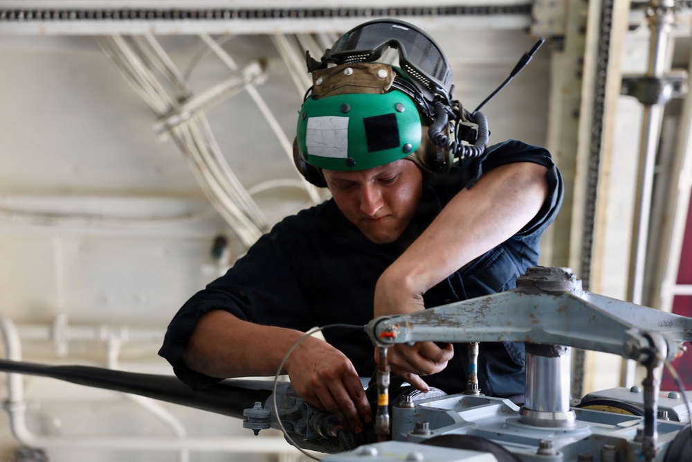 A military or naval technician wearing a green flight deck helmet with integrated hearing protection is performing maintenance on mechanical equipment, likely a helicopter rotor head or similar aircraft component. The individual is dressed in a black uniform and is working intently with both hands on metal hardware in what appears to be a hangar bay environment. The setting and equipment suggest a naval aviation maintenance context.
