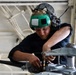 A military or naval technician wearing a green flight deck helmet with integrated hearing protection is performing maintenance on mechanical equipment, likely a helicopter rotor head or similar aircraft component. The individual is dressed in a black uniform and is working intently with both hands on metal hardware in what appears to be a hangar bay environment. The setting and equipment suggest a naval aviation maintenance context.