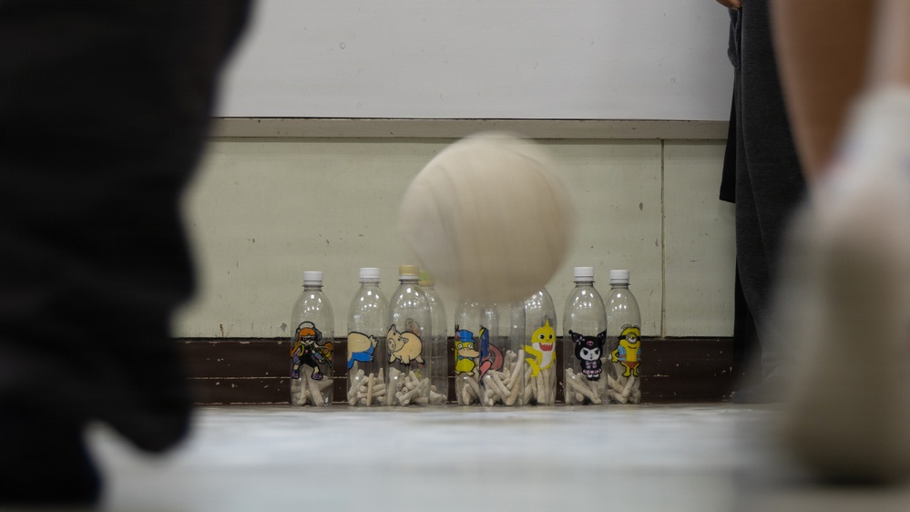 A ball is captured mid-air approaching a row of plastic bottles arranged as makeshift bowling pins. The bottles are decorated with colorful cartoon character stickers and filled with small white objects, likely serving as weights. This appears to be an improvised bowling game set up indoors.