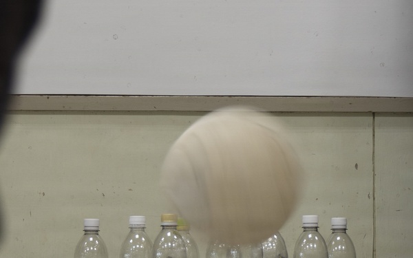 A ball is captured mid-air approaching a row of plastic bottles arranged as makeshift bowling pins. The bottles are decorated with colorful cartoon character stickers and filled with small white objects, likely serving as weights. This appears to be an improvised bowling game set up indoors.