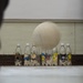 A ball is captured mid-air approaching a row of plastic bottles arranged as makeshift bowling pins. The bottles are decorated with colorful cartoon character stickers and filled with small white objects, likely serving as weights. This appears to be an improvised bowling game set up indoors.