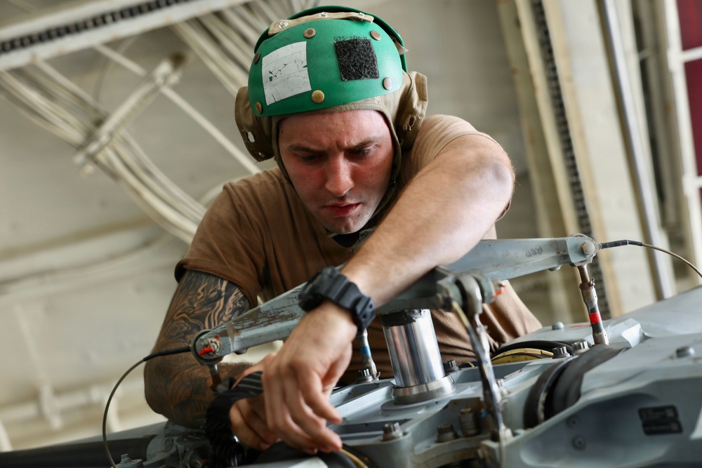 A male military or aviation technician wearing a green cranial helmet with ear protection and a boom microphone is performing maintenance or inspection work on mechanical equipment, likely aircraft components. He is leaning over the machinery with focused concentration, using his hands to manipulate cables or components. The setting appears to be an aircraft hangar or maintenance bay.