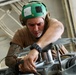 A male military or aviation technician wearing a green cranial helmet with ear protection and a boom microphone is performing maintenance or inspection work on mechanical equipment, likely aircraft components. He is leaning over the machinery with focused concentration, using his hands to manipulate cables or components. The setting appears to be an aircraft hangar or maintenance bay.