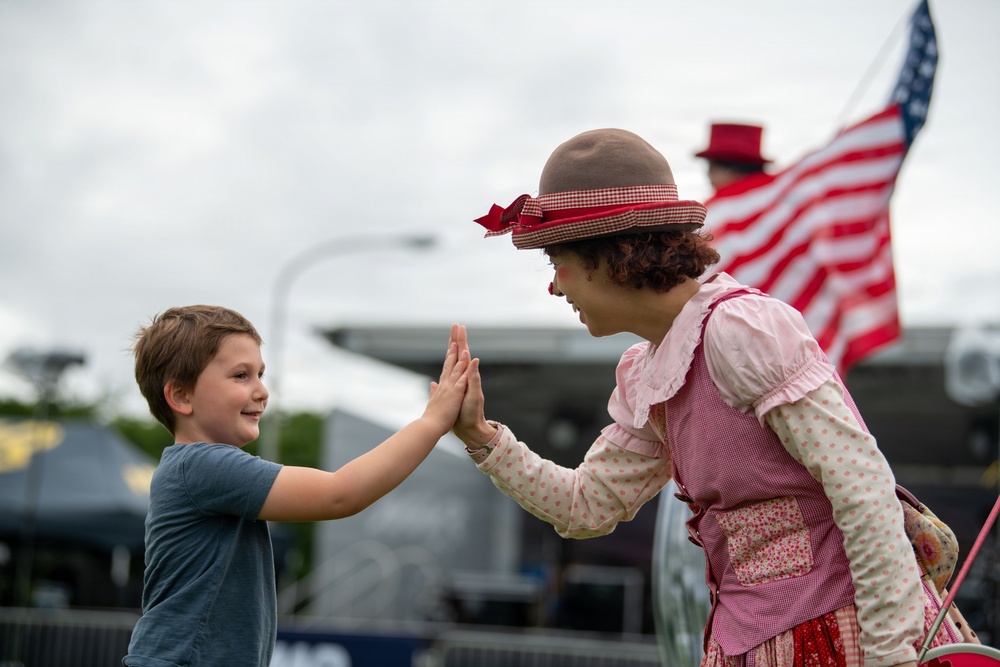 A young boy and a costumed female clown or performer are sharing a high-five at what appears to be an outdoor public event or fair. The performer is dressed in vintage-style clothing with a brown bowler hat, red gingham ribbon, and a pink floral apron. An American flag is visible in the background, suggesting a patriotic or national holiday event.