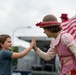 A young boy and a costumed female clown or performer are sharing a high-five at what appears to be an outdoor public event or fair. The performer is dressed in vintage-style clothing with a brown bowler hat, red gingham ribbon, and a pink floral apron. An American flag is visible in the background, suggesting a patriotic or national holiday event.
