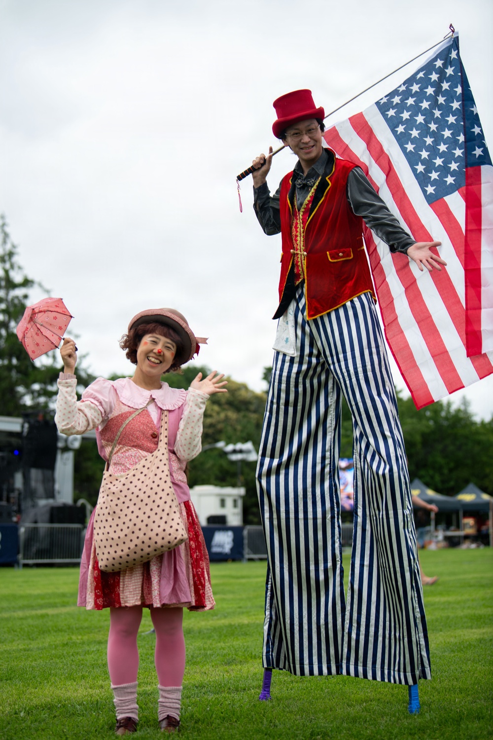 Two costumed performers pose on a grass field at an outdoor festival or event. One performer is dressed as a clown in pink attire holding a small parasol, while the other is on stilts wearing a circus ringmaster costume and carrying an American flag. The setting appears to be a public outdoor event with a stage and tents visible in the background.