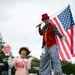 Two costumed performers pose on a grass field at an outdoor festival or event. One performer is dressed as a clown in pink attire holding a small parasol, while the other is on stilts wearing a circus ringmaster costume and carrying an American flag. The setting appears to be a public outdoor event with a stage and tents visible in the background.