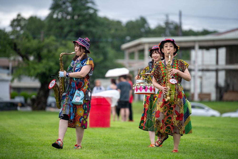 Three women dressed in vibrant, colorful patterned outfits and matching decorative hats are performing outdoors on a grassy field. Two of the women are playing saxophones while the middle performer appears to be playing a small decorative drum. They appear to be part of a street performance or outdoor festival.