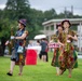 Three women dressed in vibrant, colorful patterned outfits and matching decorative hats are performing outdoors on a grassy field. Two of the women are playing saxophones while the middle performer appears to be playing a small decorative drum. They appear to be part of a street performance or outdoor festival.
