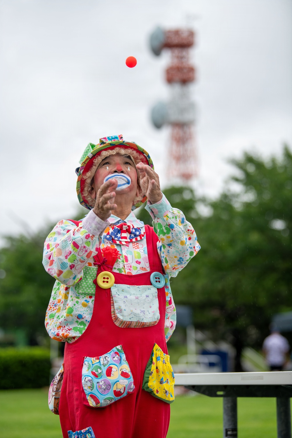 A clown in full costume is performing outdoors, juggling a red ball while looking upward. The performer wears elaborate colorful attire including a patterned hat, multicolored jacket, red overalls with large decorative buttons, and classic clown face paint with a red nose. The setting appears to be a public park or outdoor venue, with a red-and-white communications tower visible in the background.