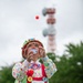 A clown in full costume is performing outdoors, juggling a red ball while looking upward. The performer wears elaborate colorful attire including a patterned hat, multicolored jacket, red overalls with large decorative buttons, and classic clown face paint with a red nose. The setting appears to be a public park or outdoor venue, with a red-and-white communications tower visible in the background.