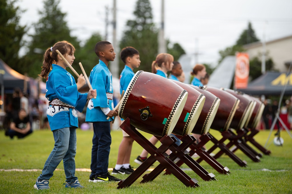 A group of children dressed in matching blue uniforms are performing taiko drumming at an outdoor cultural event or festival. They are lined up beside a row of traditional Japanese taiko drums mounted on wooden X-frame stands. The setting appears to be a grassy field with vendor tents and spectators visible in the blurred background.