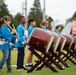 A group of children dressed in matching blue uniforms are performing taiko drumming at an outdoor cultural event or festival. They are lined up beside a row of traditional Japanese taiko drums mounted on wooden X-frame stands. The setting appears to be a grassy field with vendor tents and spectators visible in the blurred background.