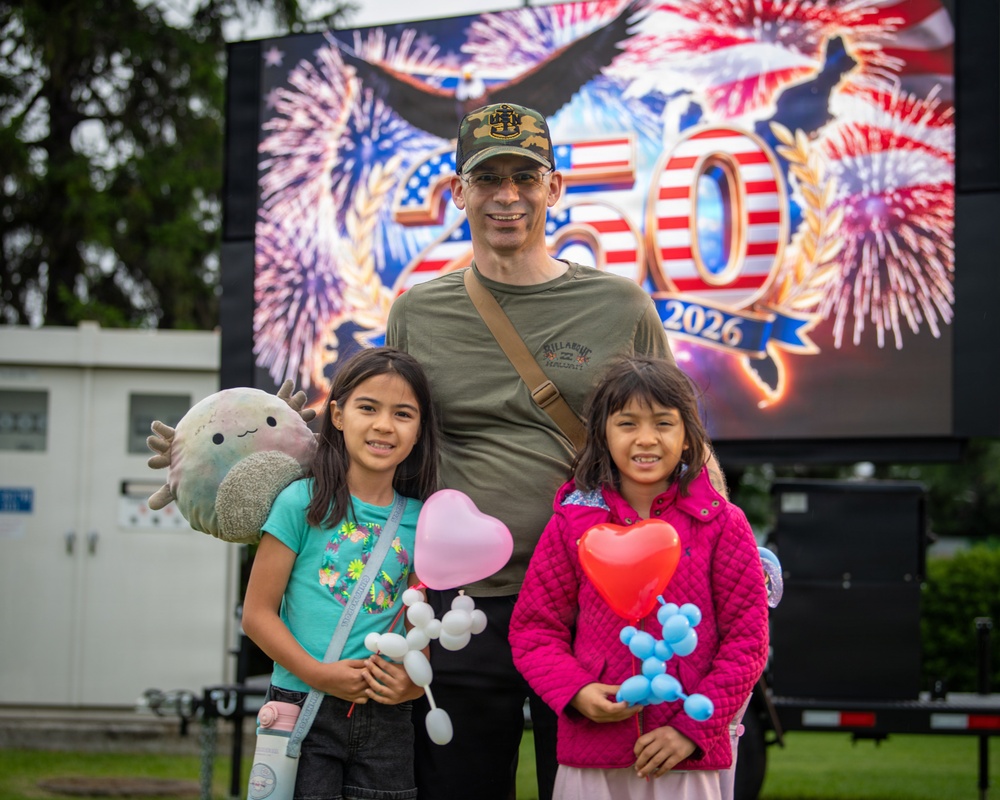 A man wearing a camouflage cap and olive green shirt poses with two young girls at what appears to be a patriotic celebration event. The girls are holding balloon animals and heart-shaped balloons, and one is carrying a Squishmallow plush toy. A large LED screen behind them displays a '250 - 2026' graphic with American flags, fireworks, and an eagle, likely referencing the United States 250th anniversary celebration.
