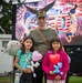 A man wearing a camouflage cap and olive green shirt poses with two young girls at what appears to be a patriotic celebration event. The girls are holding balloon animals and heart-shaped balloons, and one is carrying a Squishmallow plush toy. A large LED screen behind them displays a '250 - 2026' graphic with American flags, fireworks, and an eagle, likely referencing the United States 250th anniversary celebration.