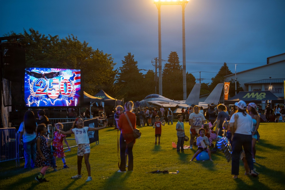 An outdoor evening patriotic celebration event, likely on a military base, featuring a large LED display showing '250 - 1776 to 2026' commemorating America's 250th anniversary. Families with children are gathered on a grass field at dusk, with a military jet aircraft (appears to be an F-15) visible in the background as a static display. Vendor tents and signage including 'MWR' (Morale, Welfare and Recreation) are visible, confirming a military installation setting.