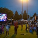 An outdoor evening patriotic celebration event, likely on a military base, featuring a large LED display showing '250 - 1776 to 2026' commemorating America's 250th anniversary. Families with children are gathered on a grass field at dusk, with a military jet aircraft (appears to be an F-15) visible in the background as a static display. Vendor tents and signage including 'MWR' (Morale, Welfare and Recreation) are visible, confirming a military installation setting.