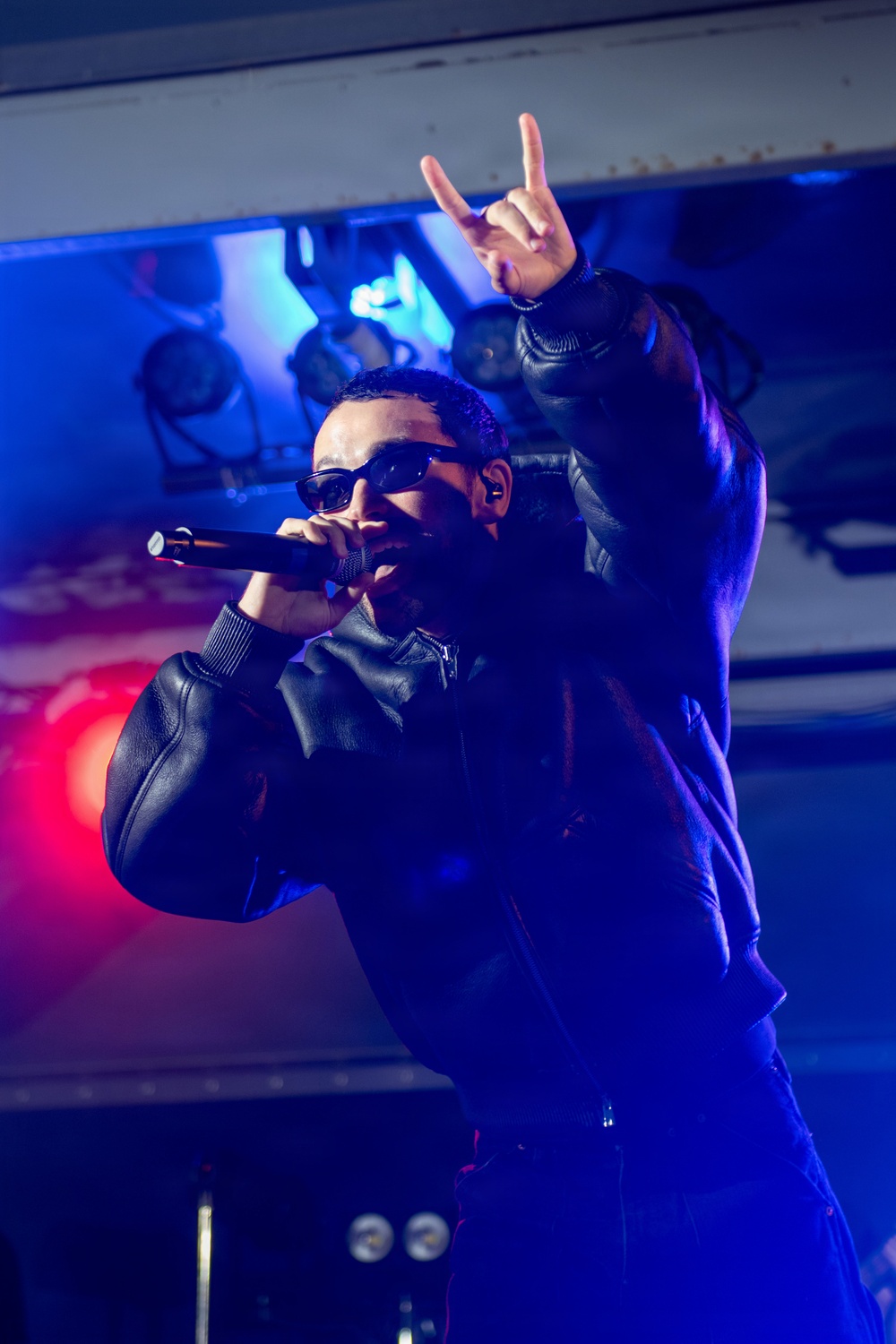 A male performer is on stage holding a microphone and making a 'rock on' or 'devil horns' hand gesture with his raised arm. He is wearing a black leather jacket and dark sunglasses under dramatic blue and red stage lighting. The setting appears to be an indoor concert or club venue.