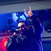 A male performer is on stage holding a microphone and making a 'rock on' or 'devil horns' hand gesture with his raised arm. He is wearing a black leather jacket and dark sunglasses under dramatic blue and red stage lighting. The setting appears to be an indoor concert or club venue.