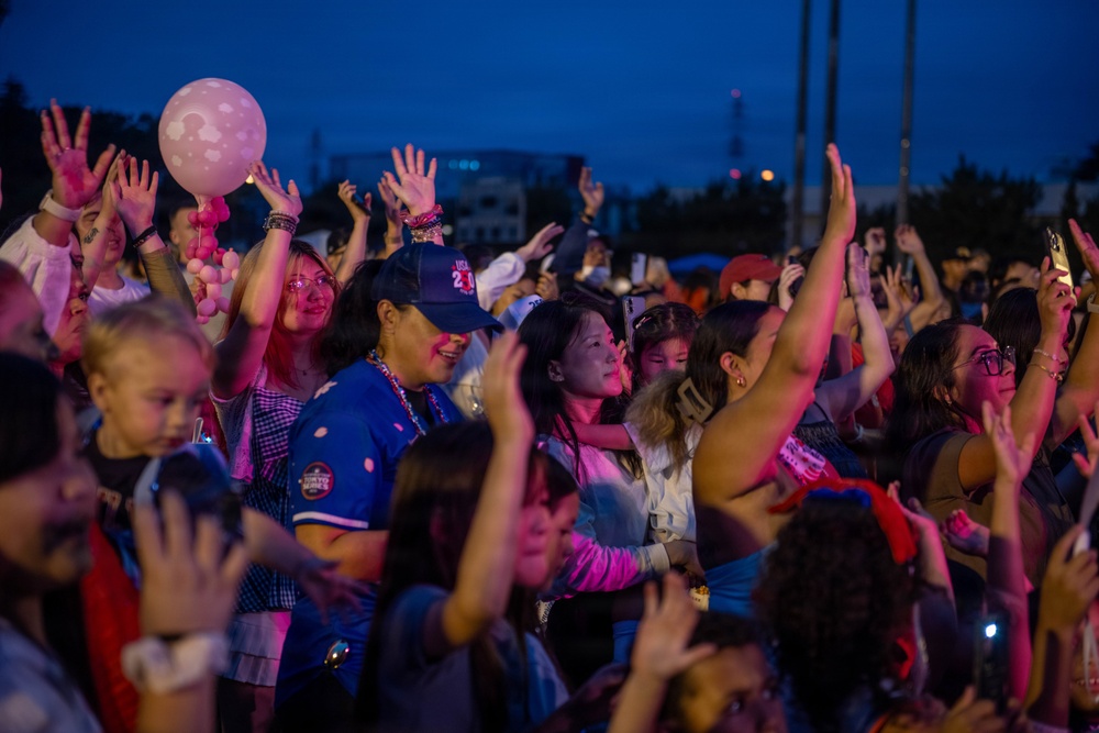A large crowd of people gathered at an outdoor evening event, likely a concert or festival, illuminated by pink and purple stage lighting. Attendees are energetically raising their hands and holding balloons, suggesting active participation in a live performance. The dusk sky and artificial lighting create a vibrant, festive atmosphere.