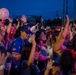 A large crowd of people gathered at an outdoor evening event, likely a concert or festival, illuminated by pink and purple stage lighting. Attendees are energetically raising their hands and holding balloons, suggesting active participation in a live performance. The dusk sky and artificial lighting create a vibrant, festive atmosphere.