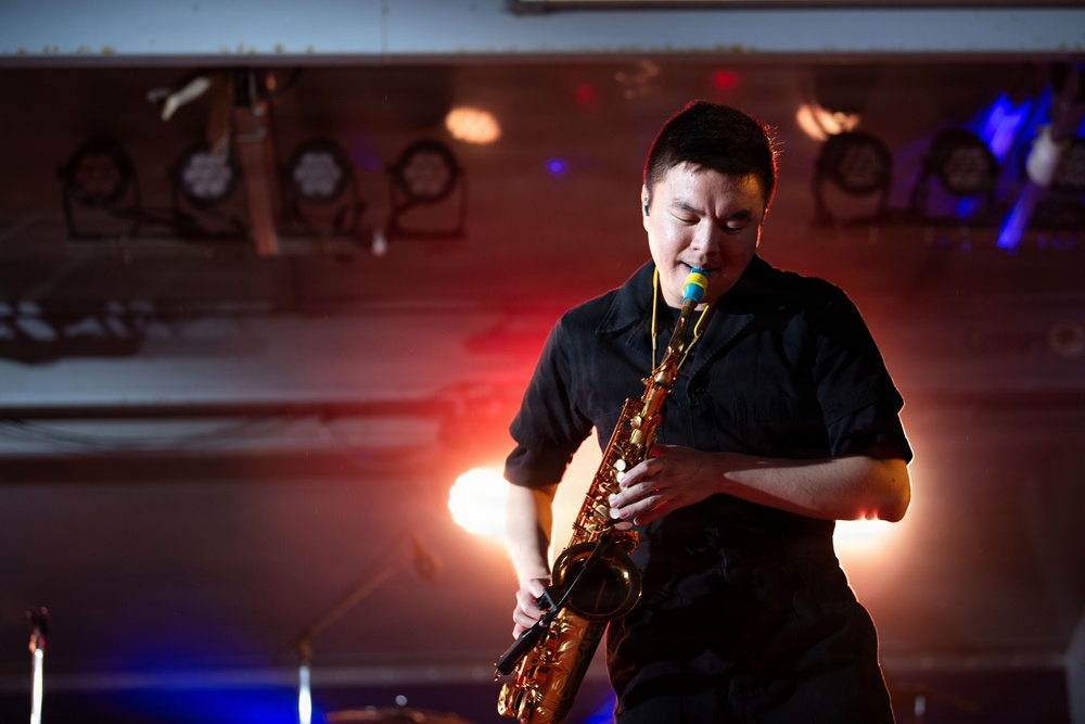 A male musician is performing on stage playing a brass saxophone under dramatic stage lighting. He is dressed in a black short-sleeve shirt and appears deeply focused on his performance with eyes downcast. The background reveals multiple stage lighting fixtures mounted on a rig, casting red, blue, and warm orange hues.