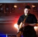 A male musician is performing on stage playing a brass saxophone under dramatic stage lighting. He is dressed in a black short-sleeve shirt and appears deeply focused on his performance with eyes downcast. The background reveals multiple stage lighting fixtures mounted on a rig, casting red, blue, and warm orange hues.