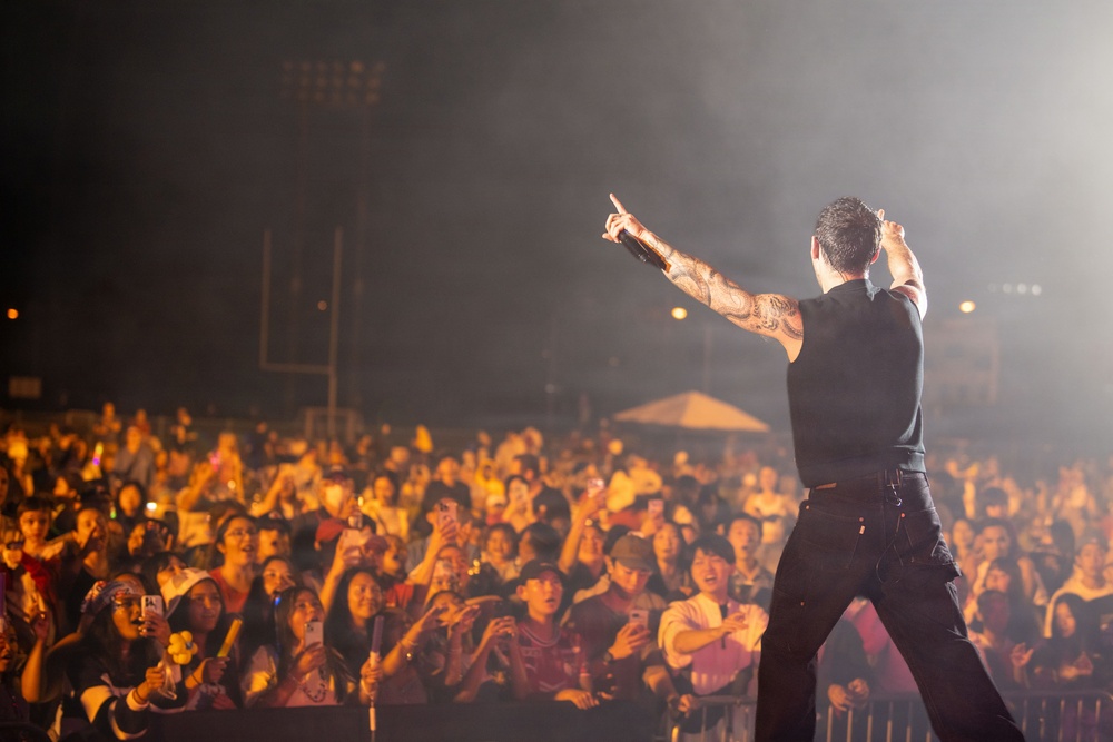 A tattooed male performer stands on stage with his back to the camera, pointing upward toward the crowd while holding a microphone. He is performing before a large, energetic audience illuminated by warm orange stage lighting. The venue appears to be an outdoor stadium or large open-air venue at night.
