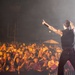 A tattooed male performer stands on stage with his back to the camera, pointing upward toward the crowd while holding a microphone. He is performing before a large, energetic audience illuminated by warm orange stage lighting. The venue appears to be an outdoor stadium or large open-air venue at night.