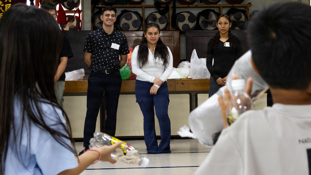 A group of people are gathered in what appears to be a cultural or community hall, with several individuals standing near a wooden bench against a wall decorated with large circular drum-like objects. Three people wearing name badges are standing at the front, appearing to be presenters or volunteers, while others in the foreground hold packaged items. The setting suggests a community event, orientation, or cultural exchange activity.