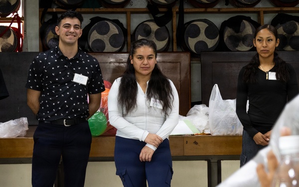 A group of people are gathered in what appears to be a cultural or community hall, with several individuals standing near a wooden bench against a wall decorated with large circular drum-like objects. Three people wearing name badges are standing at the front, appearing to be presenters or volunteers, while others in the foreground hold packaged items. The setting suggests a community event, orientation, or cultural exchange activity.