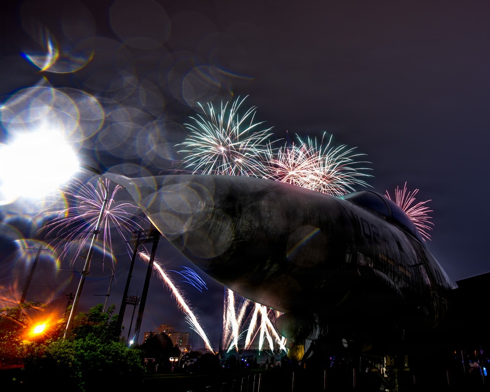 A nighttime long-exposure photograph captures a fireworks display over what appears to be a submarine or large naval vessel on display, likely at a museum or memorial site. Multiple firework bursts in teal, pink, white, and purple illuminate the dark sky above the dark hull of the vessel. The image features significant lens flare and bokeh effects from a nearby bright light source, adding to the dramatic visual composition.
