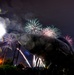 A nighttime long-exposure photograph captures a fireworks display over what appears to be a submarine or large naval vessel on display, likely at a museum or memorial site. Multiple firework bursts in teal, pink, white, and purple illuminate the dark sky above the dark hull of the vessel. The image features significant lens flare and bokeh effects from a nearby bright light source, adding to the dramatic visual composition.