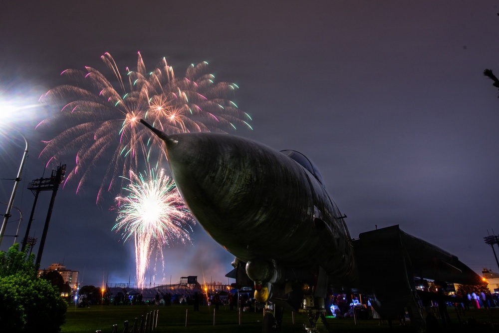 A nighttime outdoor event featuring a fireworks display bursting in pink, green, and white colors over a large dark-colored aircraft or military jet on static display. A crowd of spectators is gathered around the exhibit in what appears to be an airfield or aviation museum grounds. Stadium-style floodlights and city buildings are visible in the background.