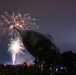A nighttime outdoor event featuring a fireworks display bursting in pink, green, and white colors over a large dark-colored aircraft or military jet on static display. A crowd of spectators is gathered around the exhibit in what appears to be an airfield or aviation museum grounds. Stadium-style floodlights and city buildings are visible in the background.