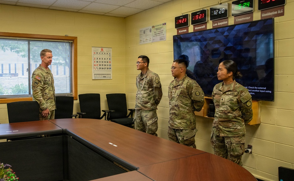 Four U.S. Army soldiers in OCP (Operational Camouflage Pattern) uniforms are gathered in a military briefing or operations room. One senior soldier on the left appears to be addressing or briefing three other soldiers standing at attention. The room contains multiple time zone clocks on the wall, a large display monitor, and a conference table.