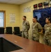 Four U.S. Army soldiers in OCP (Operational Camouflage Pattern) uniforms are gathered in a military briefing or operations room. One senior soldier on the left appears to be addressing or briefing three other soldiers standing at attention. The room contains multiple time zone clocks on the wall, a large display monitor, and a conference table.
