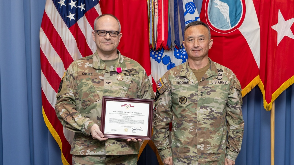 Two U.S. Army soldiers pose for a formal military award ceremony photograph. The soldier on the left, identified by name tape as 'CHENEY' and holding a framed Legion of Merit certificate, is wearing the Legion of Merit medal on his uniform. The soldier on the right, identified as 'DOOGHAN,' appears to be a Major General based on the two-star insignia visible on his uniform.