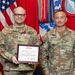 Two U.S. Army soldiers pose for a formal military award ceremony photograph. The soldier on the left, identified by name tape as 'CHENEY' and holding a framed Legion of Merit certificate, is wearing the Legion of Merit medal on his uniform. The soldier on the right, identified as 'DOOGHAN,' appears to be a Major General based on the two-star insignia visible on his uniform.