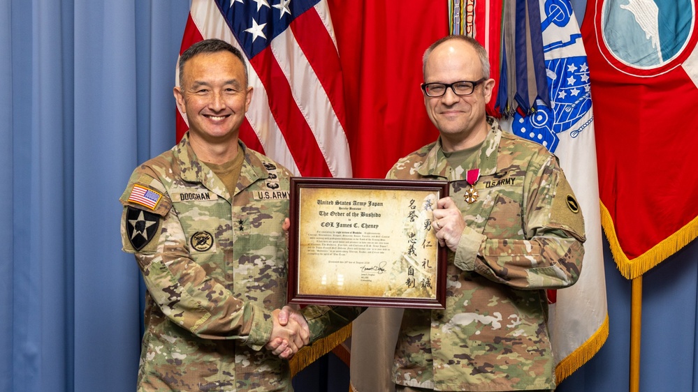 Two U.S. Army soldiers pose together holding a framed certificate from United States Army Japan titled 'The Order of the Bushido,' awarded to COL James C. Cheney. The soldier on the left, identified by his name tape as 'DOOGHAN,' appears to be a general officer based on his rank insignia, and is presenting the award. The recipient on the right is wearing a medal on his uniform, indicating this is a formal military awards ceremony.