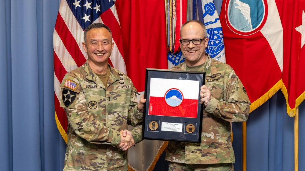 Two U.S. Army soldiers in OCP (Operational Camouflage Pattern) uniforms are posing for a formal photo while shaking hands and holding a framed military award or commemorative plaque. The soldier on the left, identified by name tape as 'DOOHAN,' appears to be a general officer based on visible rank insignia, while the soldier on the right is identified as 'U.S. ARMY' with the name partially visible as 'Cheney' on the plaque. The ceremony appears to take place in a formal military setting with multiple flags displayed in the background.