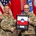 Two U.S. Army soldiers in OCP (Operational Camouflage Pattern) uniforms are posing for a formal photo while shaking hands and holding a framed military award or commemorative plaque. The soldier on the left, identified by name tape as 'DOOHAN,' appears to be a general officer based on visible rank insignia, while the soldier on the right is identified as 'U.S. ARMY' with the name partially visible as 'Cheney' on the plaque. The ceremony appears to take place in a formal military setting with multiple flags displayed in the background.